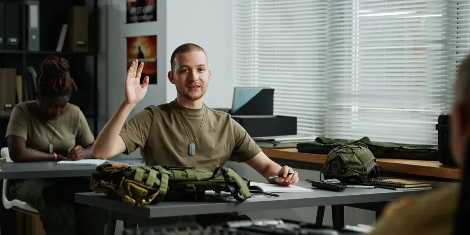 Young male student raising hand and looking at teacher at lesson while asking him question after lecture about military training